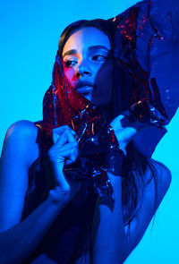 Confident female model wrapped in plastic film standing in provocative posture in studio with blue neon illumination and looking away