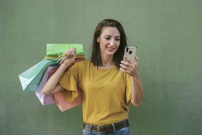 Smiling woman using smart phone holding shopping bags against wall
