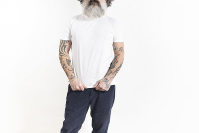 Man with tattooed arms and beard in white t-shirt and blue pants on white background.