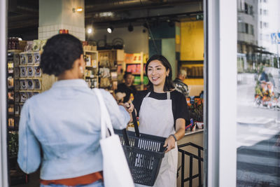 Smiling sales clerk giving shopping basket to female customer arriving at convenience store