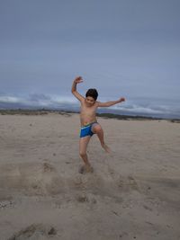 Shirtless boy jumping against cloudy sky at beach