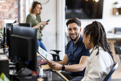 Happy businessman holding charts and graphs with colleague in front of computer