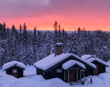 Snow covered houses and trees on field against sky during sunset