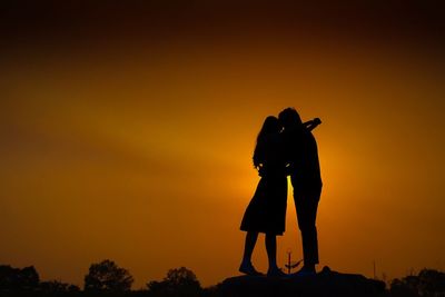Silhouette romantic couple embracing while standing on field against orange sky during sunset