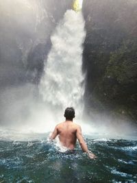Rear view of shirtless man looking at waterfall in river