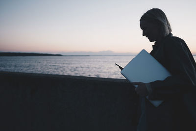 Woman using mobile phone and holding laptop while walking by lake during sunset