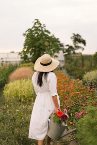 Beautiful brunette girl in a white dress and hat with watering can watering flowers