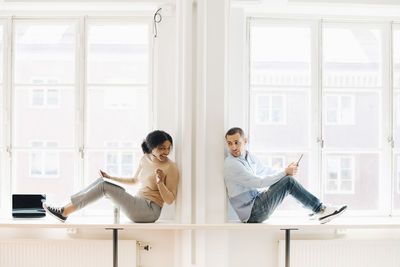 Colleagues talking while sitting on desk by window in creative office