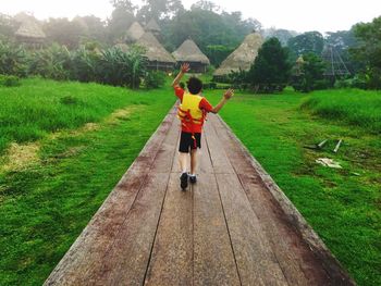 Rear view of boy with arms raised walking on boardwalk amidst grassy field