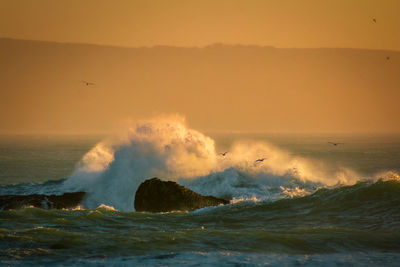 Waves splashing on sea against sky during sunset