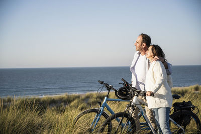 Smiling couple with bicycles looking at view while standing on beach against clear sky during weekend