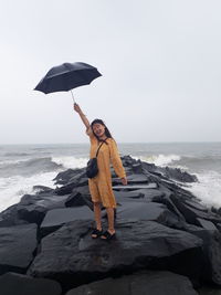 Cheerful woman holding umbrella while standing on groyne against clear sky