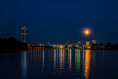 Illuminated buildings by sea against sky at night