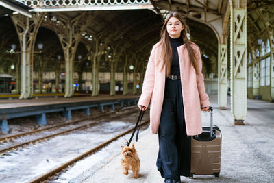 Travel concept. at station, young tourist with dog goes and drags suitcase