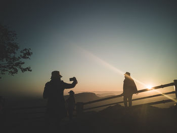 Silhouette man photographing friend against sky during sunset