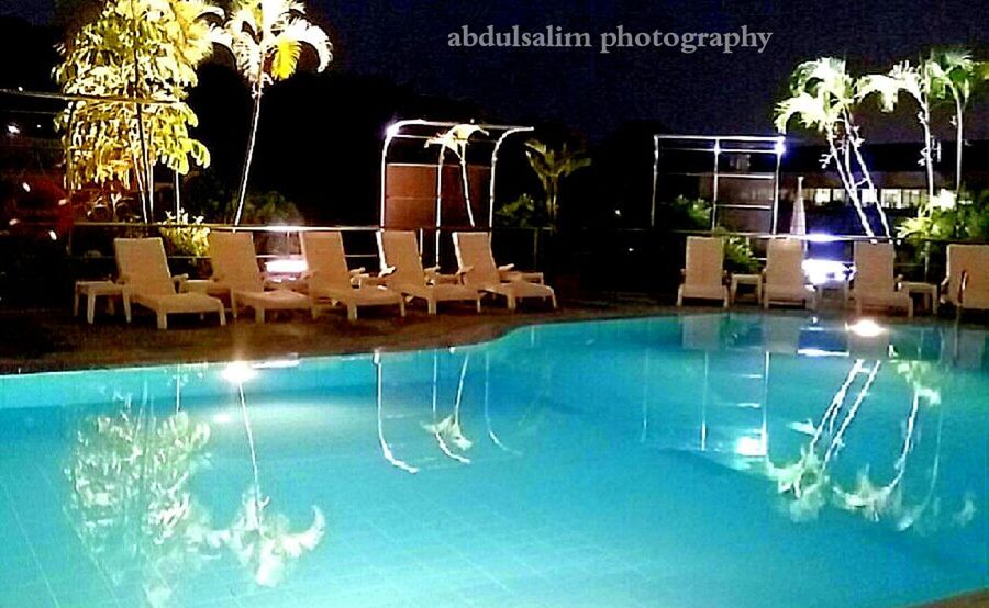 swimming pool, water, palm tree, built structure, architecture, blue, building exterior, reflection, tree, tourist resort, potted plant, sunlight, growth, plant, illuminated, window, waterfront, chair, no people, glass - material