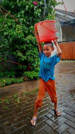 Full length of boy carrying a big bucket on his head to gather rain drops just for fun.