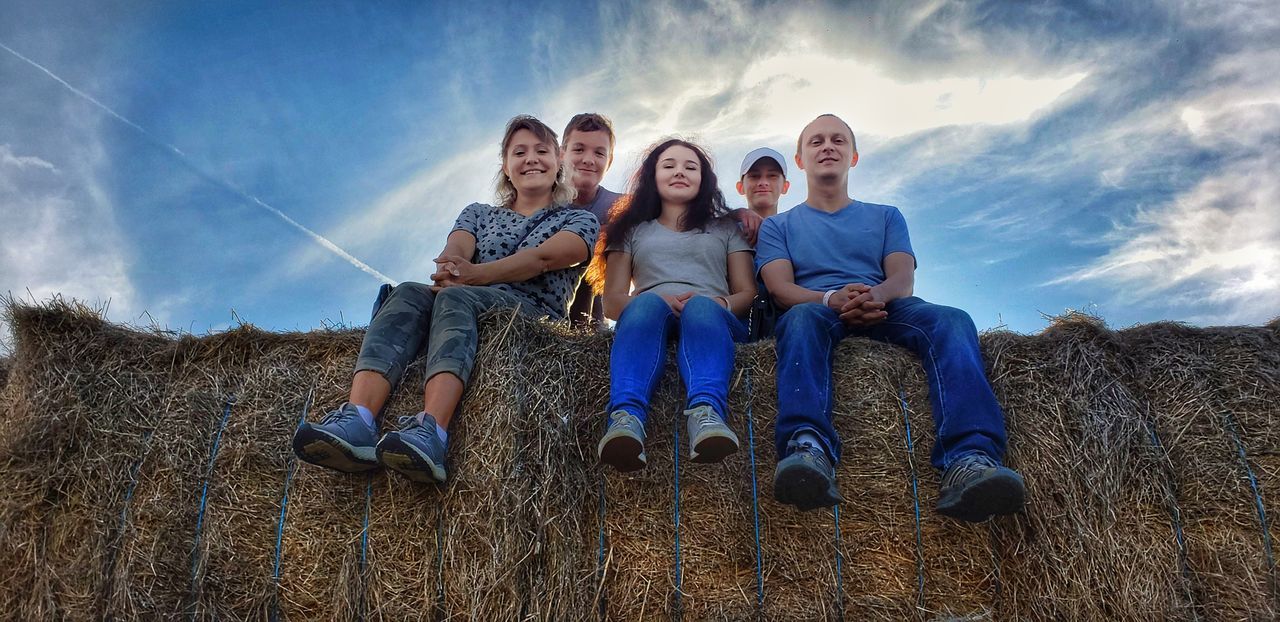 togetherness, sky, looking at camera, family, group of people, real people, portrait, front view, leisure activity, cloud - sky, happiness, casual clothing, bonding, emotion, sitting, nature, field, child, lifestyles, land, sister, outdoors, positive emotion, teenager