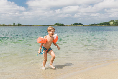 Full length of shirtless boy wearing water wings running on beach against cloudy sky