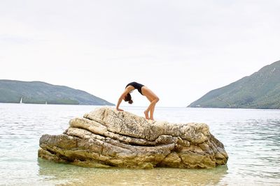 Rear view of woman in swimsuit doing yoga on rock by sea against sky