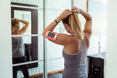 Woman in sport clothing tying ponytail while looking in mirror at home