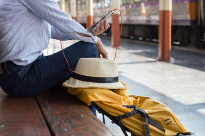 Midsection of woman listening music over mobile phone while sitting on bench at station