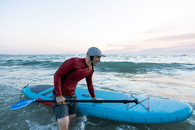 Side view of male surfer in wetsuit and hat carrying paddle board and entering water to surf on seashore