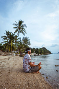 Man sitting on rock by palm trees against sky