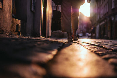 Low section of woman carrying shopping bag while walking on footpath during sunset