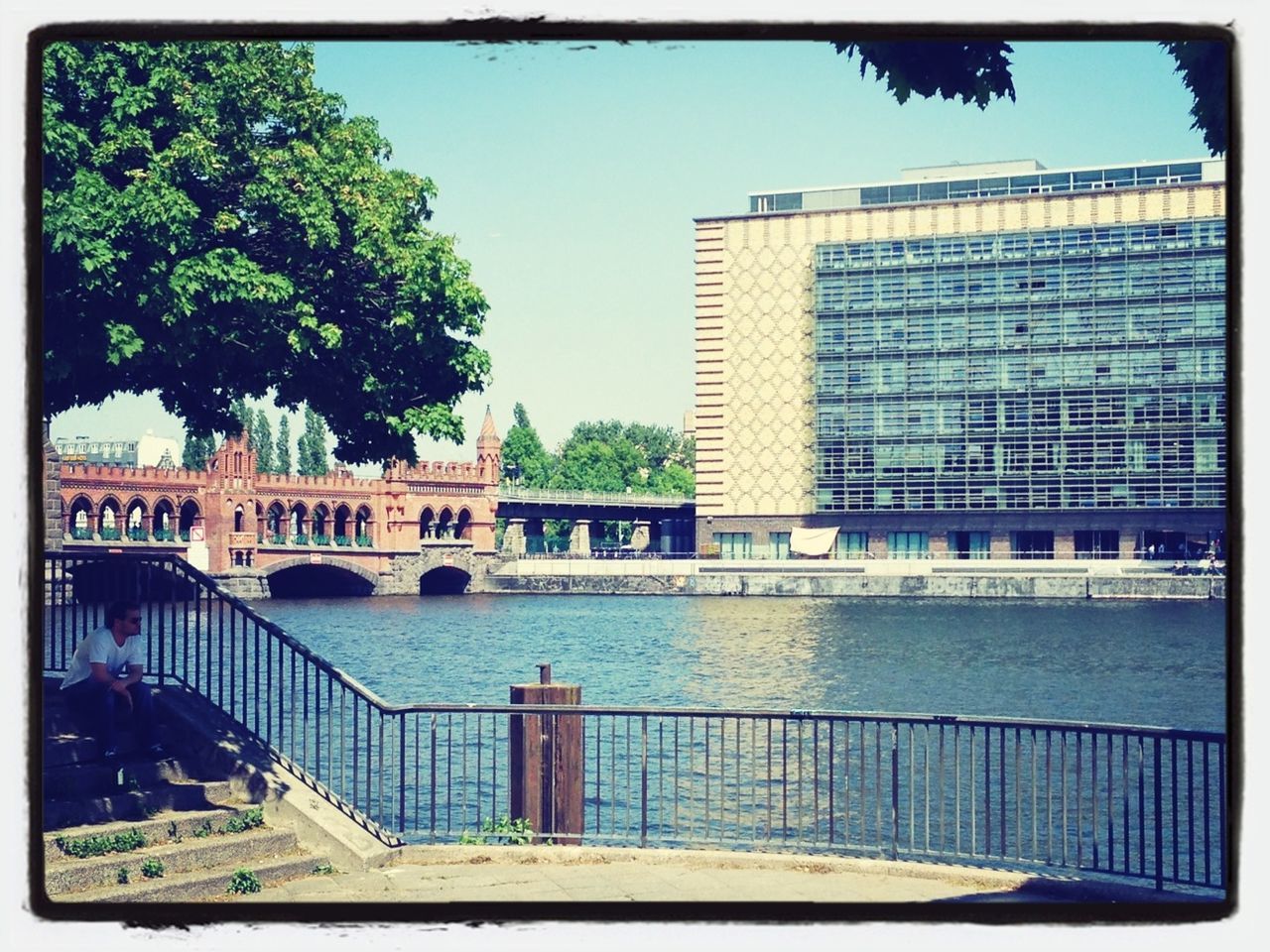 transfer print, building exterior, built structure, architecture, tree, auto post production filter, water, railing, city, sunlight, day, river, outdoors, building, clear sky, sky, reflection, growth, blue, no people