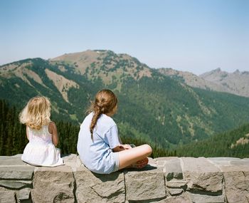 Rear view of girls sitting on stone wall against mountain