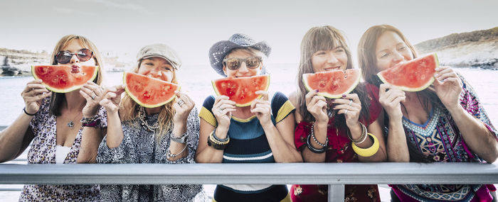 Portrait of friends eating watermelon while standing against sky