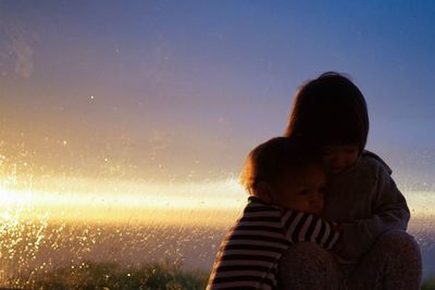 Siblings embracing against glass window during sunset