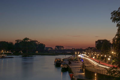 Illuminated city by river against sky at sunset