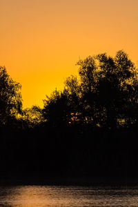 Silhouette trees by lake against orange sky