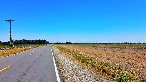 Country road amidst field against clear blue sky