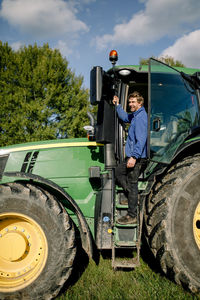 Happy mature farmer standing on tractor at farm under cloudy sky