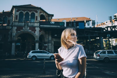 Young woman looking over shoulder while holding mobile phone on street in city