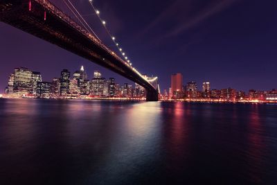 Illuminated bridge over river by buildings against sky at night