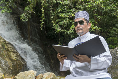 Man reading book while sitting against waterfall
