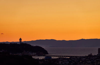 Silhouette buildings by sea against orange sky