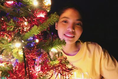 Portrait of young woman smiling by illuminated christmas tree in darkroom