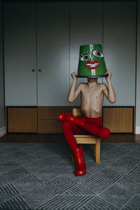 Shirtless man covering face with bucket at home