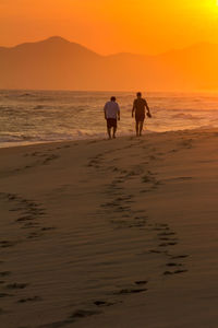 Rear view of men walking on beach against sky during sunset