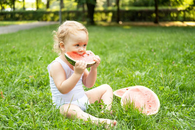 Funny smiling kid boy in white bodysuit eating watermelon at green lawn