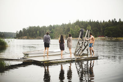 Friends wrapped in towels standing on jetty over lake against clear sky