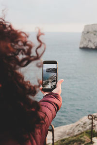 Rear view of woman photographing with mobile phone against sea