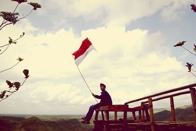 Smiling man with indonesia flag sitting on observation point over mountain against sky
