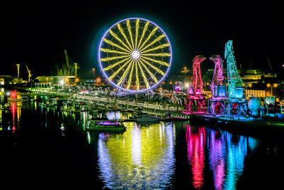 Illuminated ferris wheel by river against sky at night