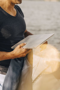 Midsection of man holding laptop while standing against lake